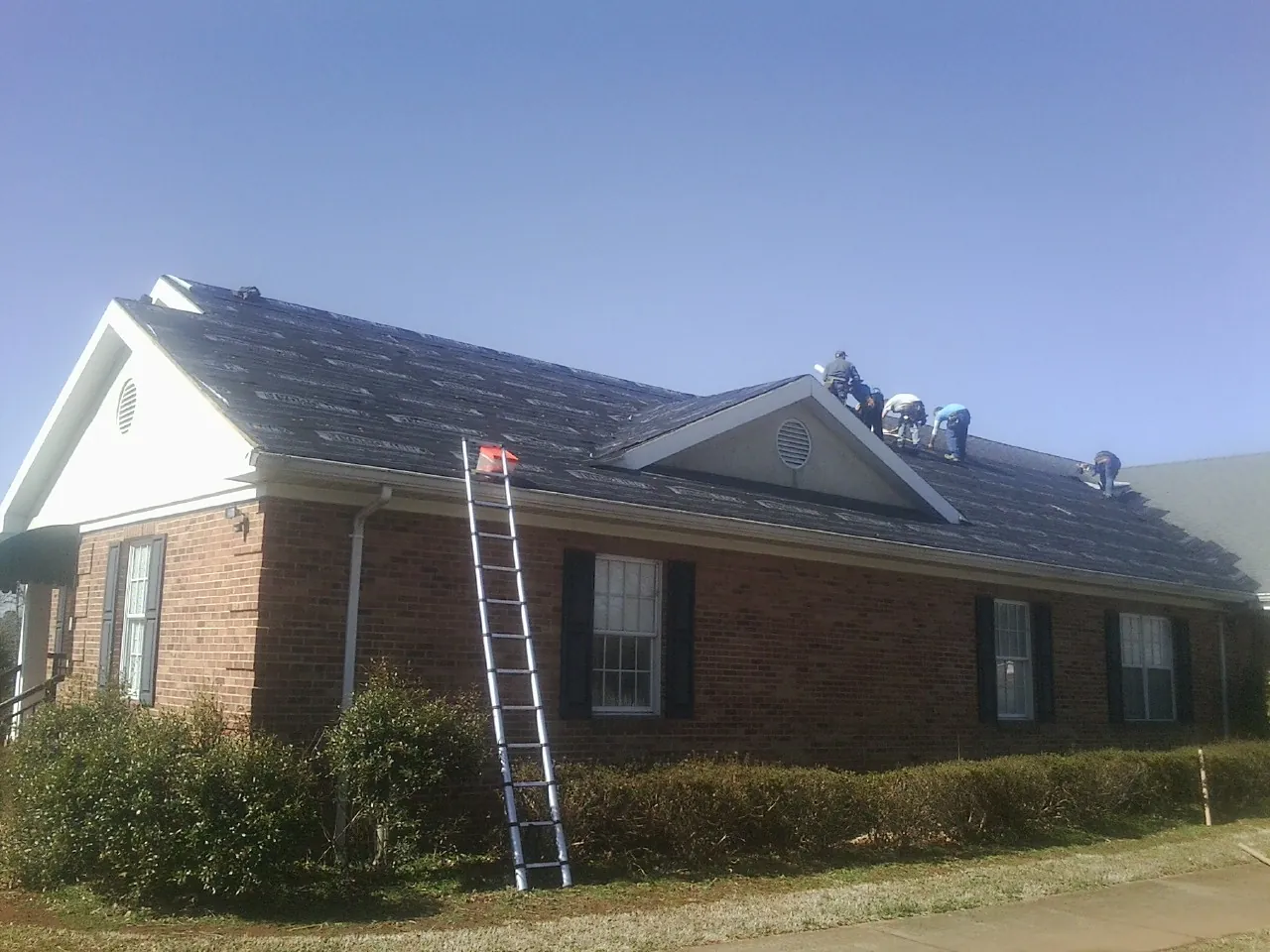 Crew tearing shingles off a brick ranch roof with a ladder set — Metal Roof Repair work by our crew in Granby